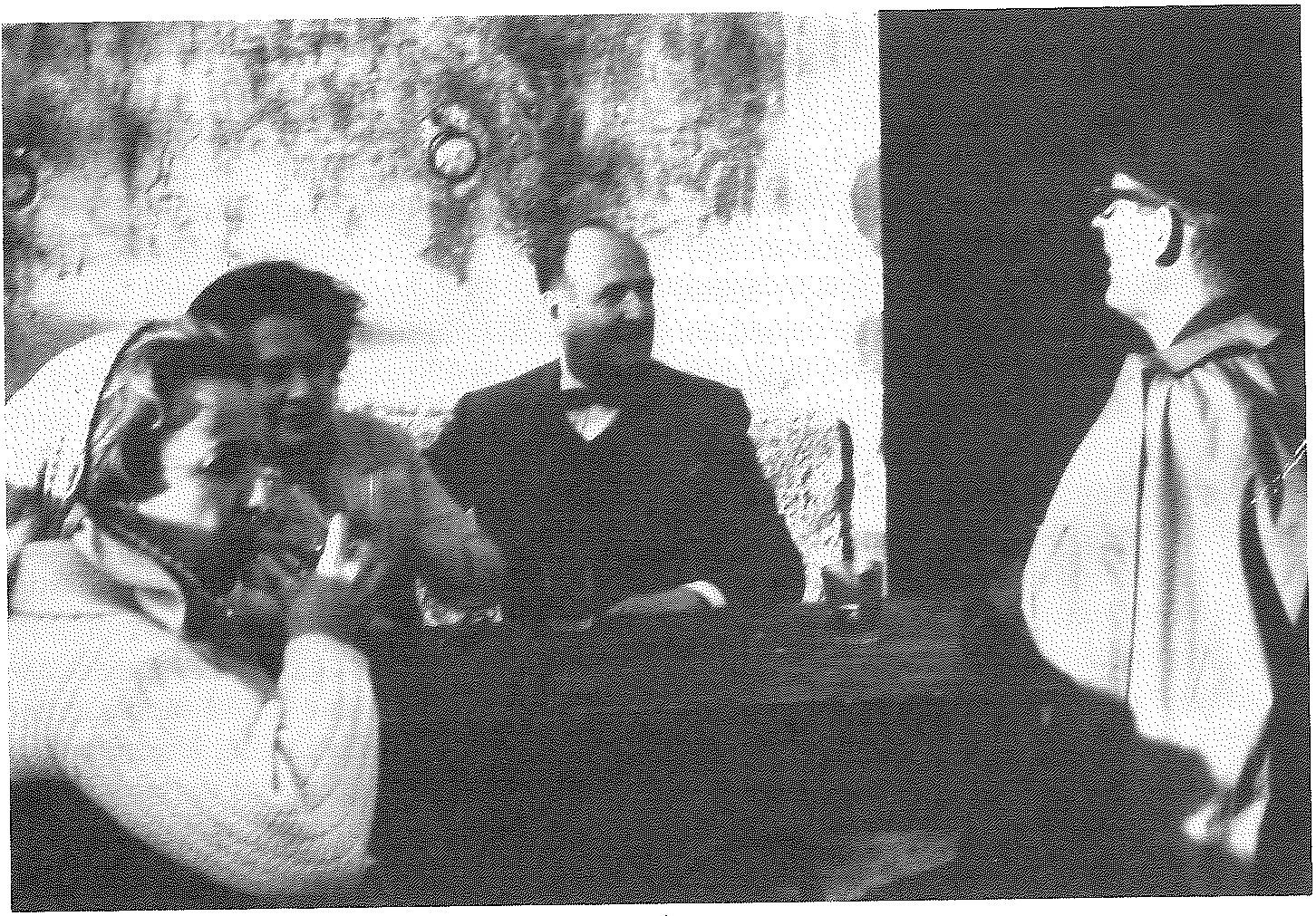 Two couples sitting and talking in a sunlit room, a figure standing in the doorway at the right.