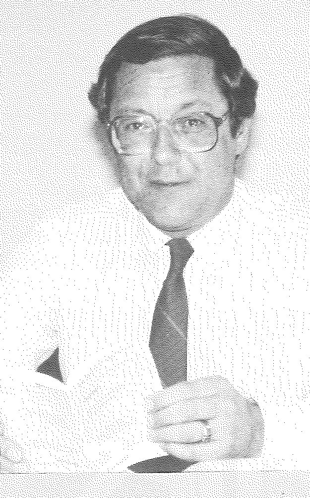 A bespectacled man with a moustache, in a dark suit and tie, seated behind a desk.