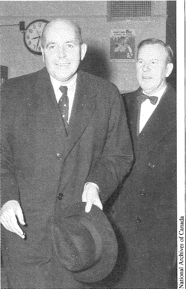 Two men in heavy overcoats stand side by side indoors, the taller holding a hat, a wall clock behind them.