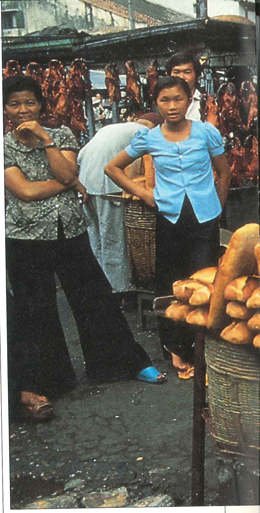 Two women waiting at a street stall hung with roast ducks and meats, beside baskets of baguettes