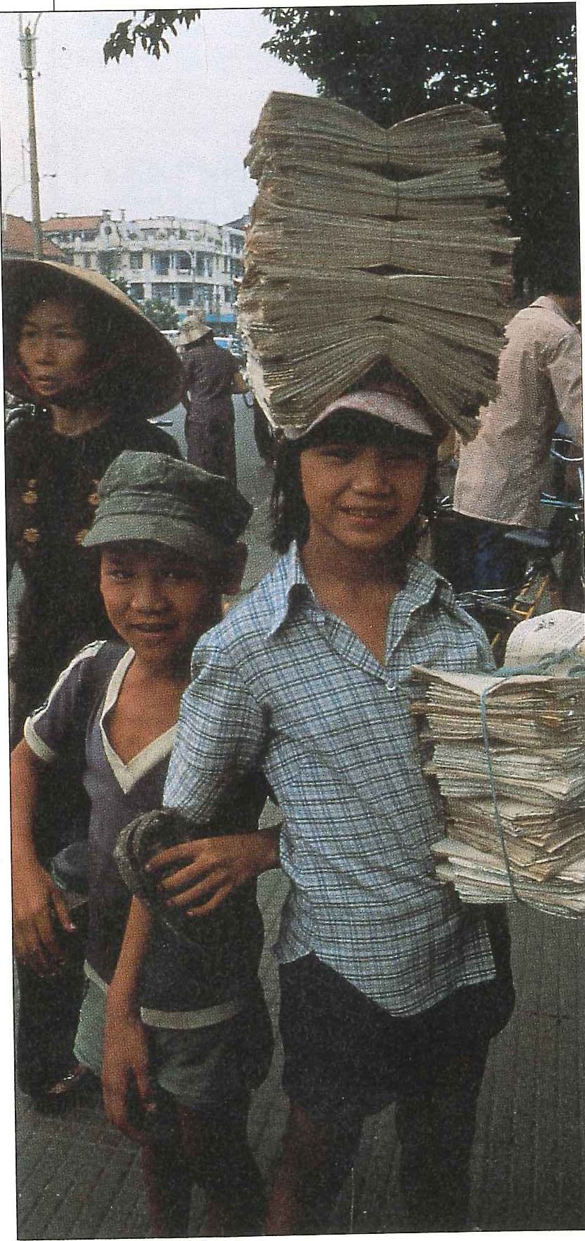 Two boys in a busy street, one carrying a tall stack of newspapers balanced on his head