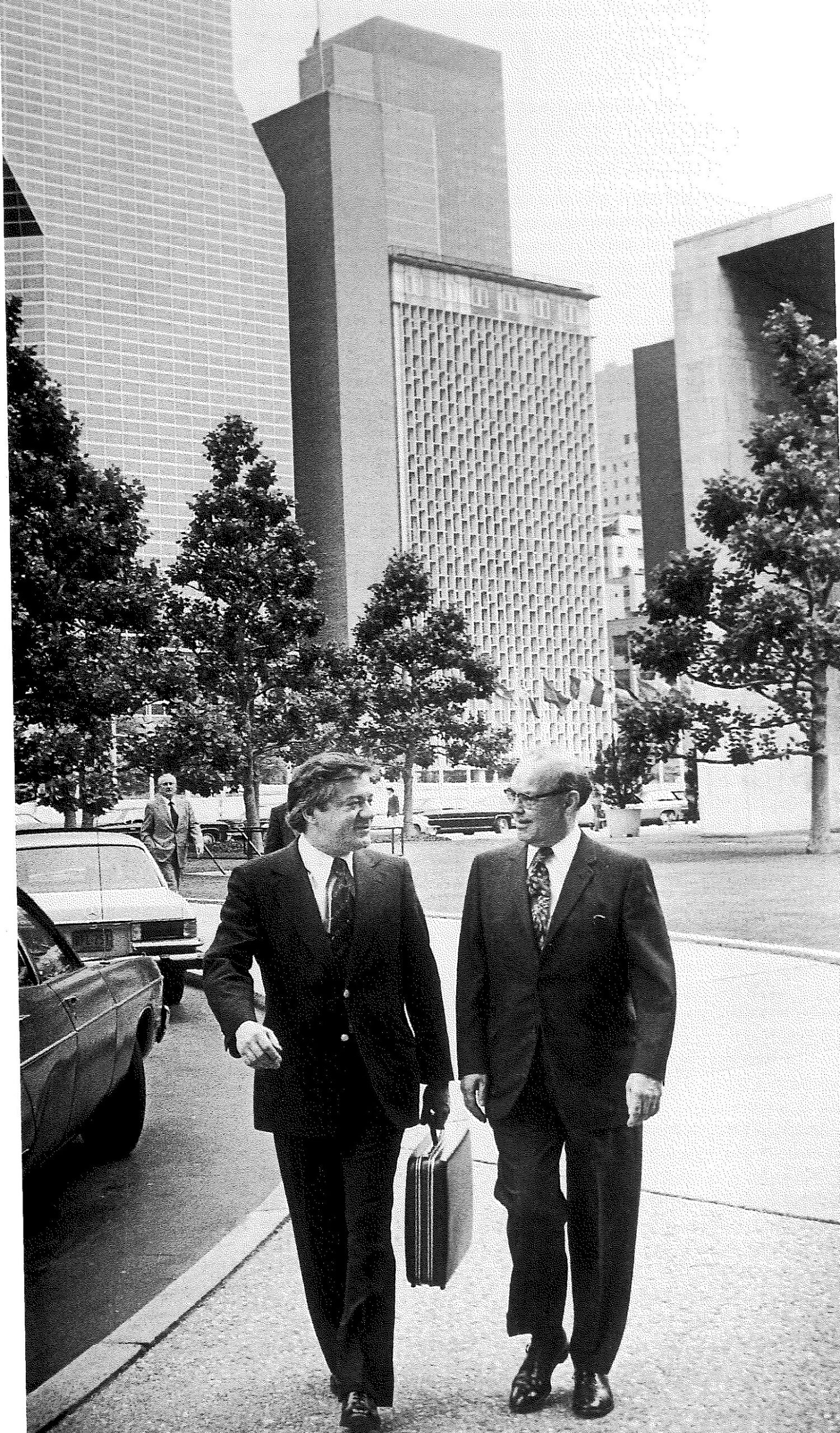 Two men in suits walk along a tree-lined street past parked cars, with office towers behind them