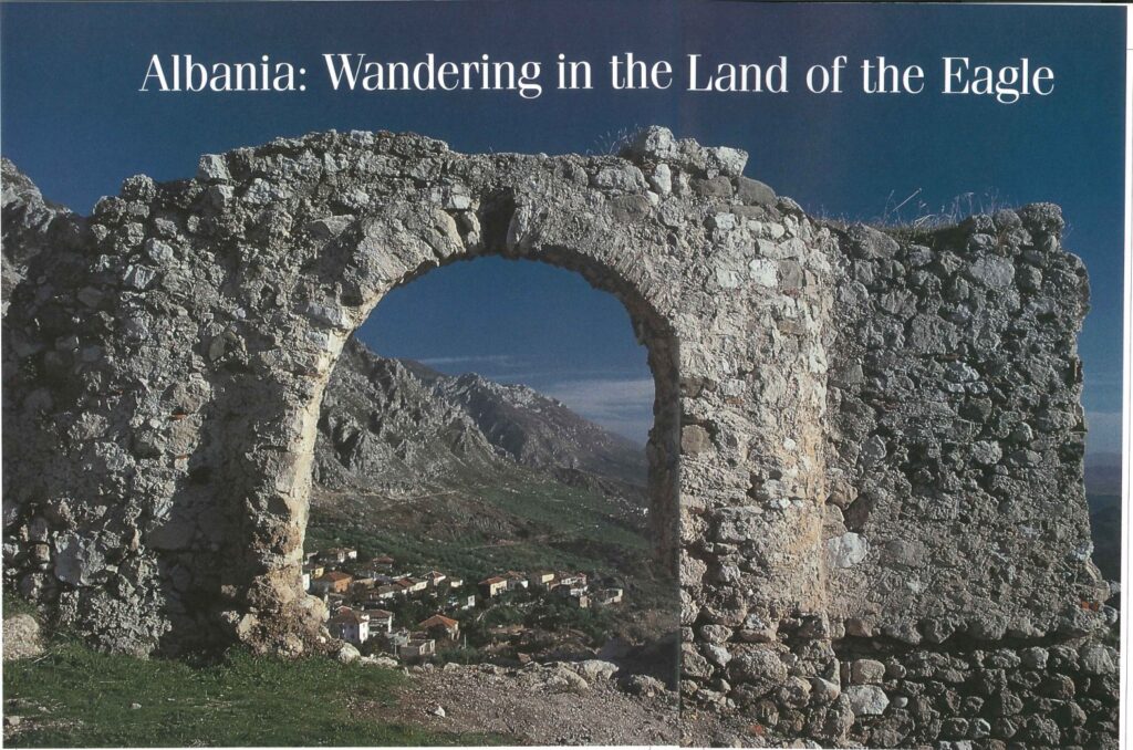 A ruined stone arch on a hilltop, with a village and bare mountains beyond