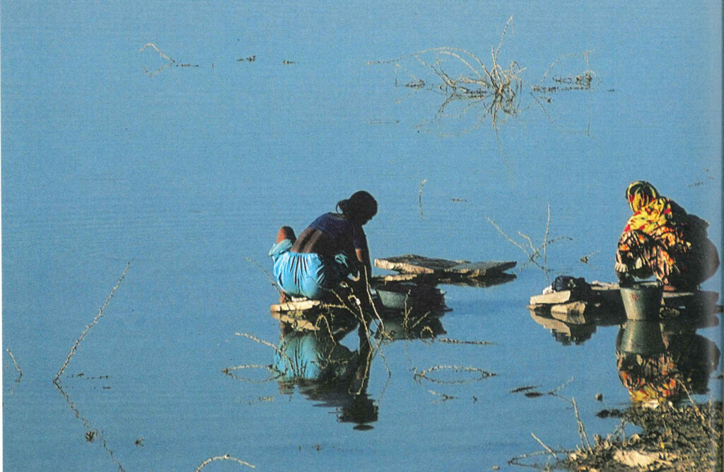 Two women crouched at the edge of a wide river, washing clothes and filling pots