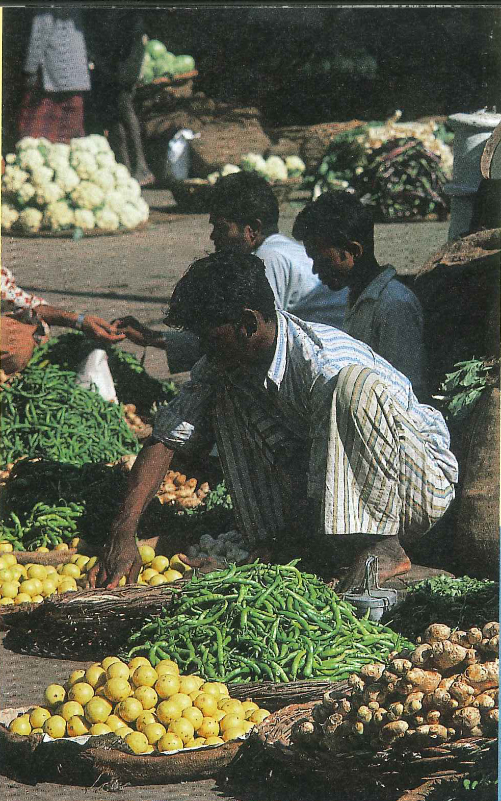 Vendors and customers around baskets of lemons, green chillies, ginger and cauliflower at an open-air market