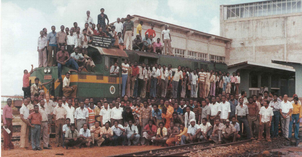 A crowd of railway workers standing on and around a diesel locomotive outside a workshop