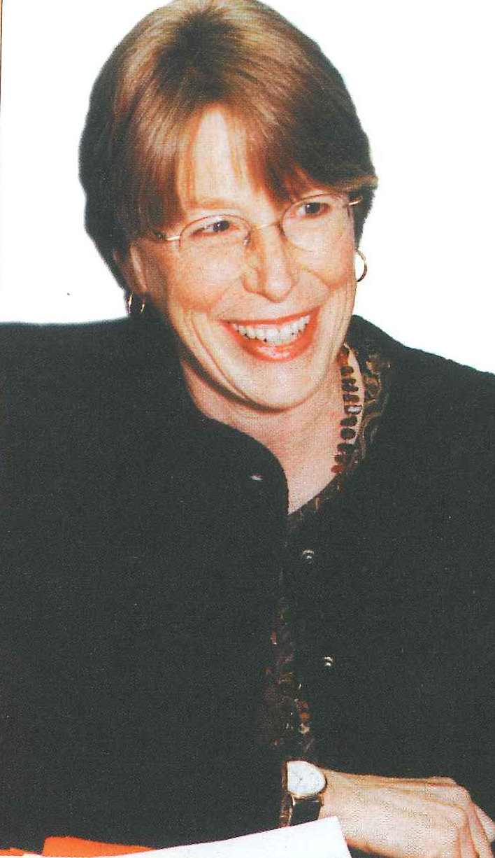 A smiling woman in a dark jacket seated at a desk, her hands on a sheaf of papers