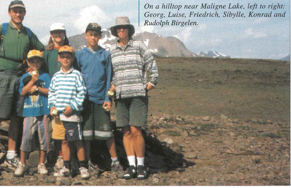 Six people of three generations standing on a bare hilltop, mountains behind them