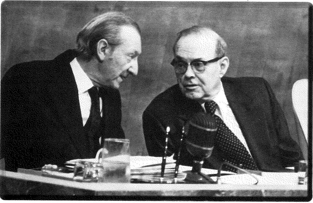 Two men in suits seated side by side at a table set with microphones