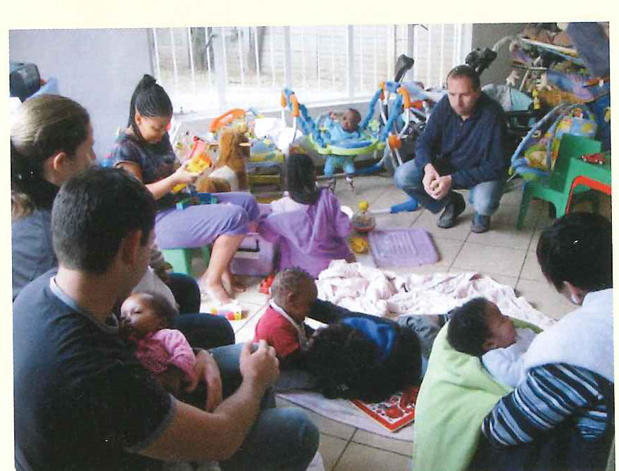 Three volunteers sit on the floor of a bright playroom among half a dozen infants and toddlers.