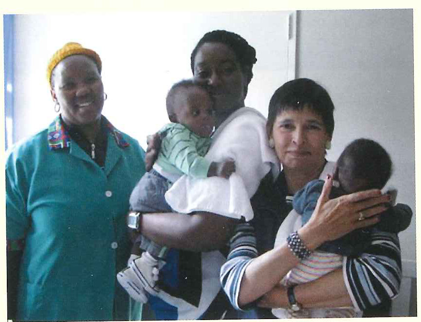 Two women stand side by side indoors, each holding a baby.