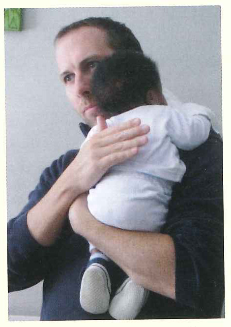 A man cradles a baby in a white outfit against his shoulder.