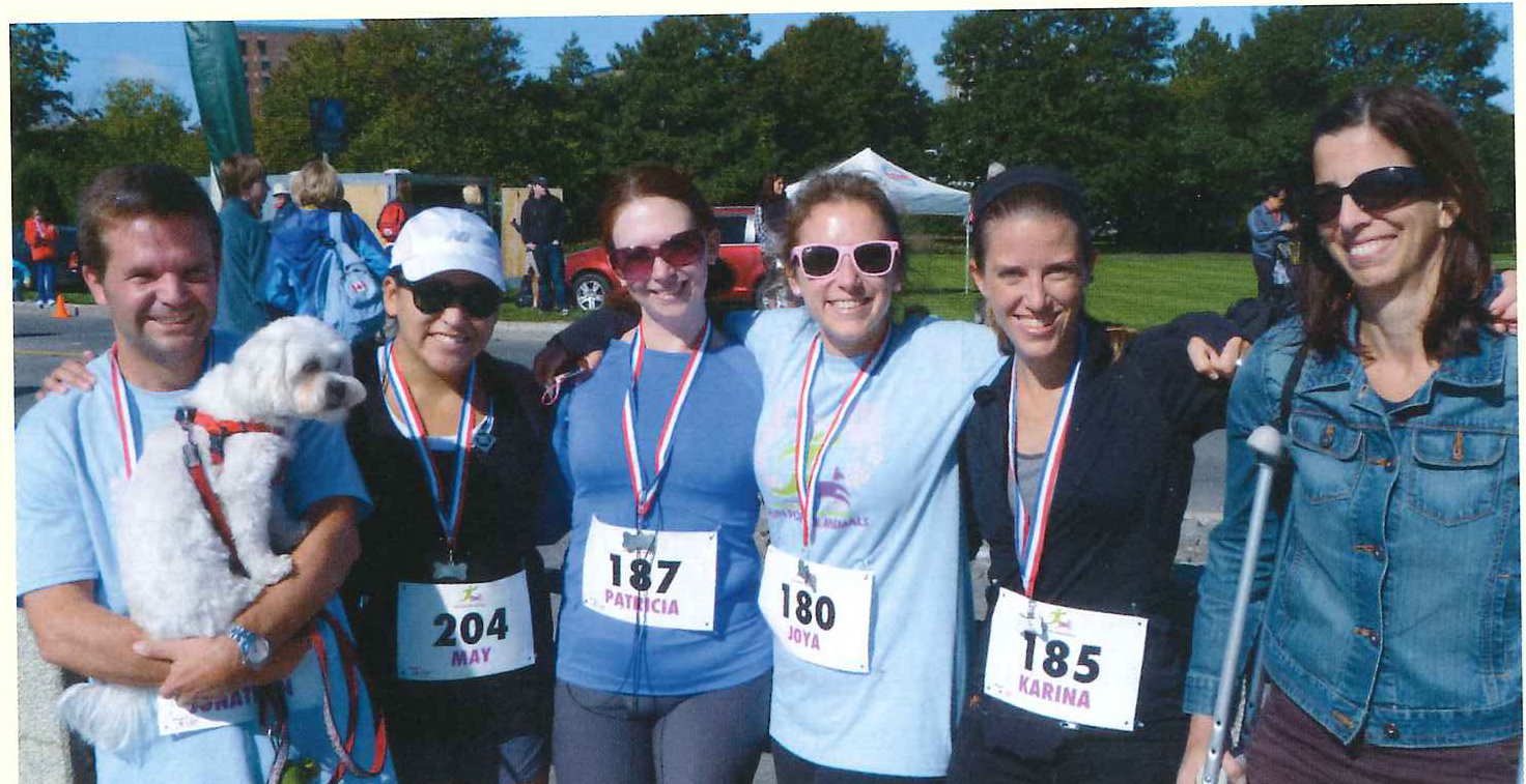 Six runners in matching blue shirts and numbered race bibs pose on the grass with a small white dog.