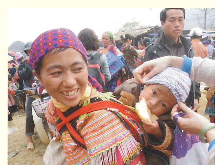 A young woman in embroidered Hmong dress smiles in a crowded market while a hand settles a knitted hat on a baby beside her.
