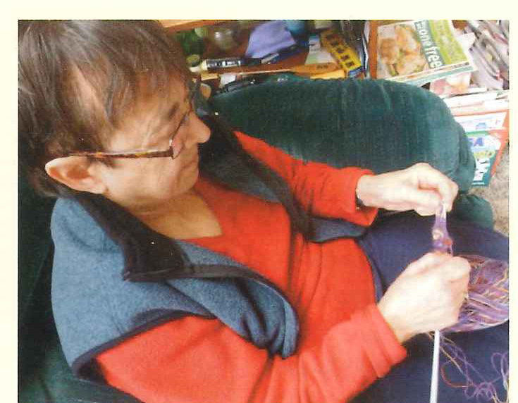 An older woman in a red and blue top sits knitting in an armchair at home.