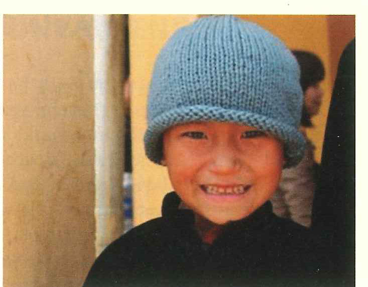 A grinning boy in a grey knitted hat stands against a plain wall.