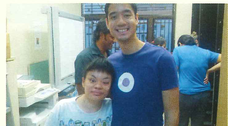A volunteer in a blue T-shirt stands smiling beside a student in a workroom.