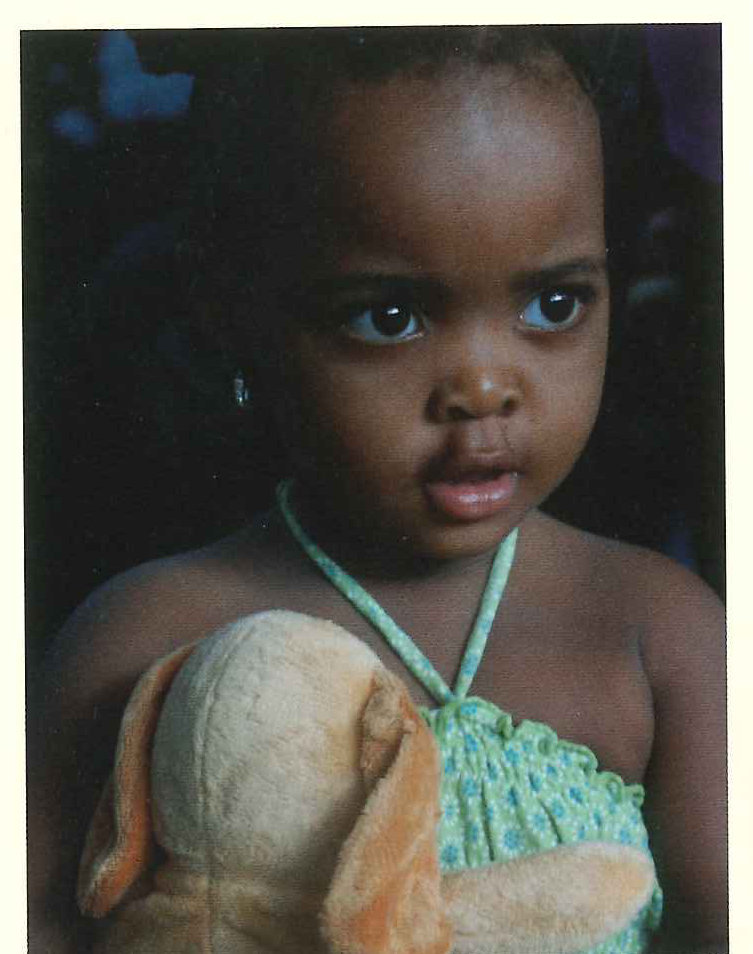 A small girl in a green dress holds a loaf of bread and looks straight at the camera.