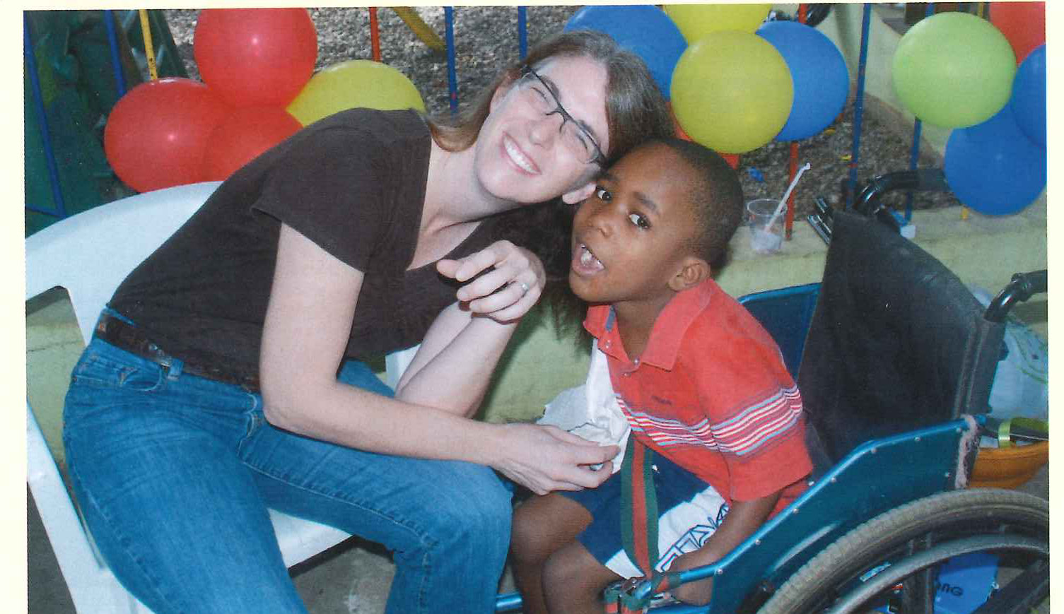 A woman crouches to hug a young boy sitting in a wheelchair beneath a cluster of balloons.