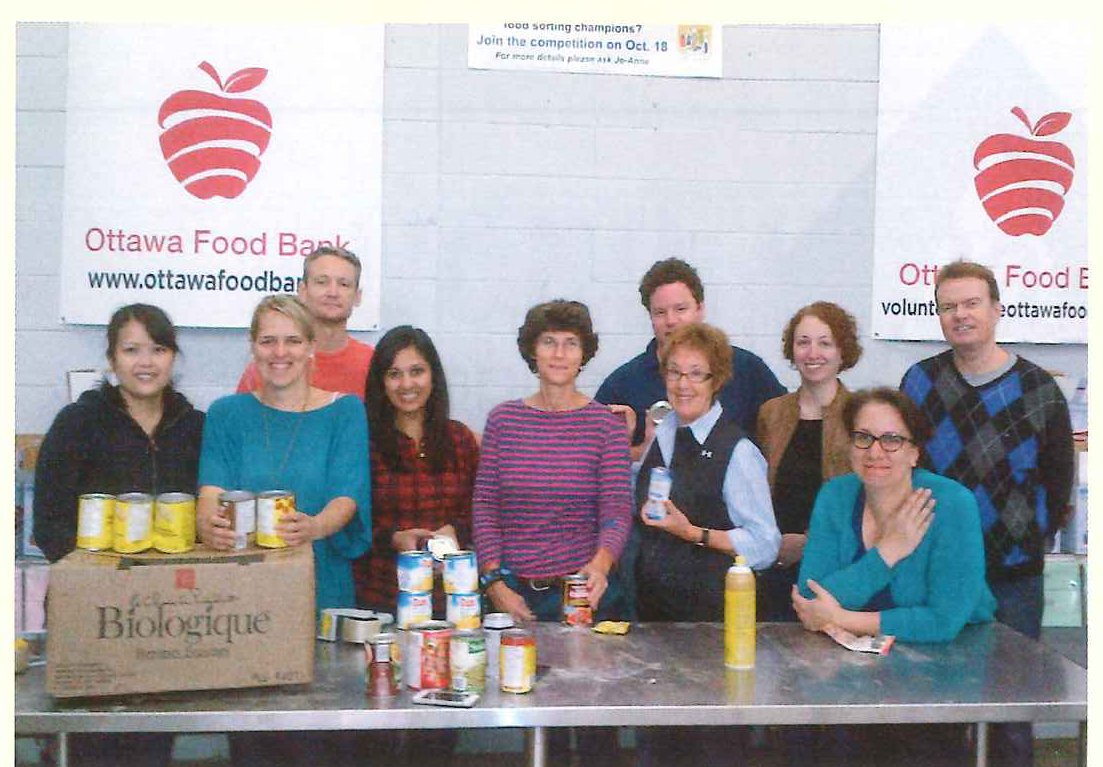 Un groupe de bénévoles pose derrière des tables chargées de conserves, sous l’enseigne de la Banque d’alimentation d’Ottawa.