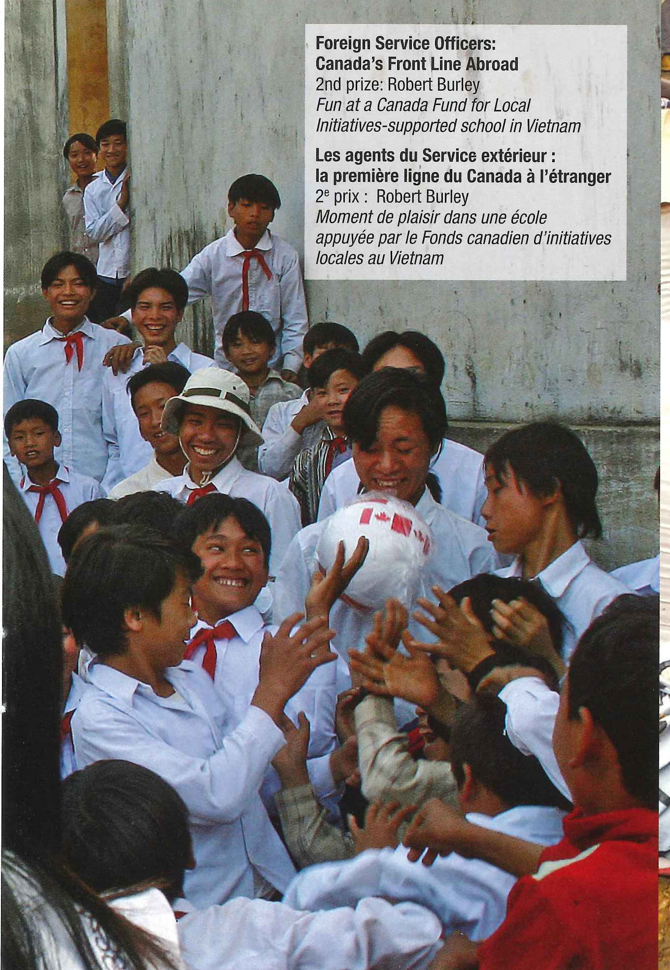 Schoolchildren in white shirts and red scarves crowd around a ball marked with a maple leaf // Des écoliers en chemise blanche et foulard rouge se pressent autour d’un ballon frappé d’une feuille d’érable