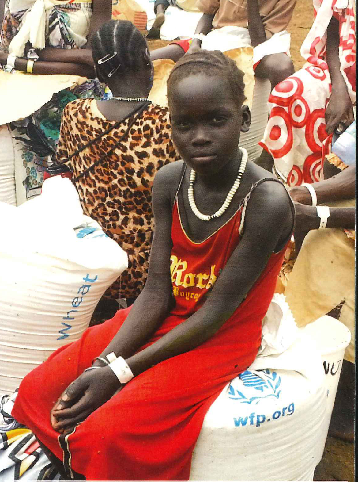 A girl in a red dress sits on sacks of food aid in a camp // Une fillette en robe rouge est assise sur des sacs d’aide alimentaire dans un camp