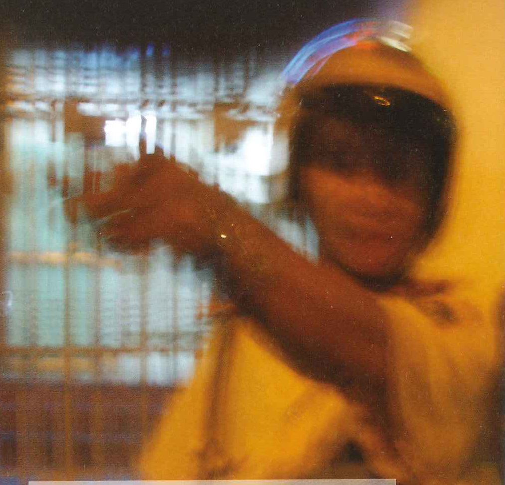 A blurred figure in a helmet reaches out in front of a barred window // Une silhouette floue casquée tend le bras devant une fenêtre grillagée
