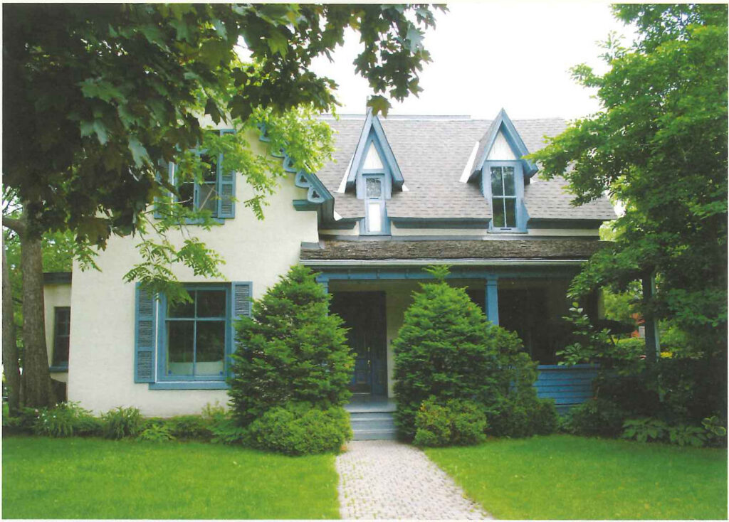 A two-storey clapboard house with a steep gabled roof behind a lawn