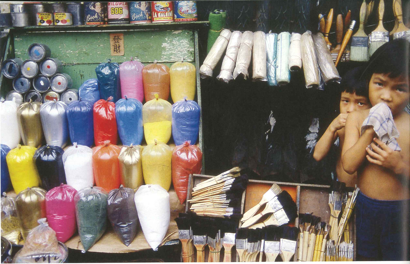A street stall hung with bundles of coloured paper and brushes, two boys at the right